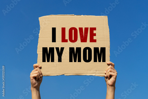 I love my mom text on box paper held by 2 hands with isolated blue sky background. This message board can be used as business concept about loving mother.