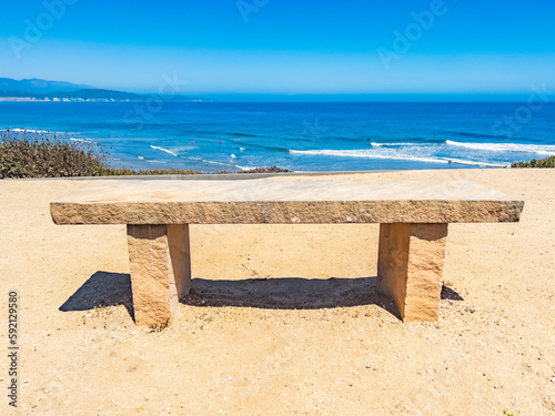 Peaceful Scene of Empty Sandstone Bench Overlooking a Serene Blue Ocean Waves Crashing Ashore in the horizon