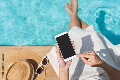 Asian woman sitting by the pool while using mobile phone. Young traveler female working on cellphone during her summer vacation trip. Technology and lifestyle concept. Copy space, top view