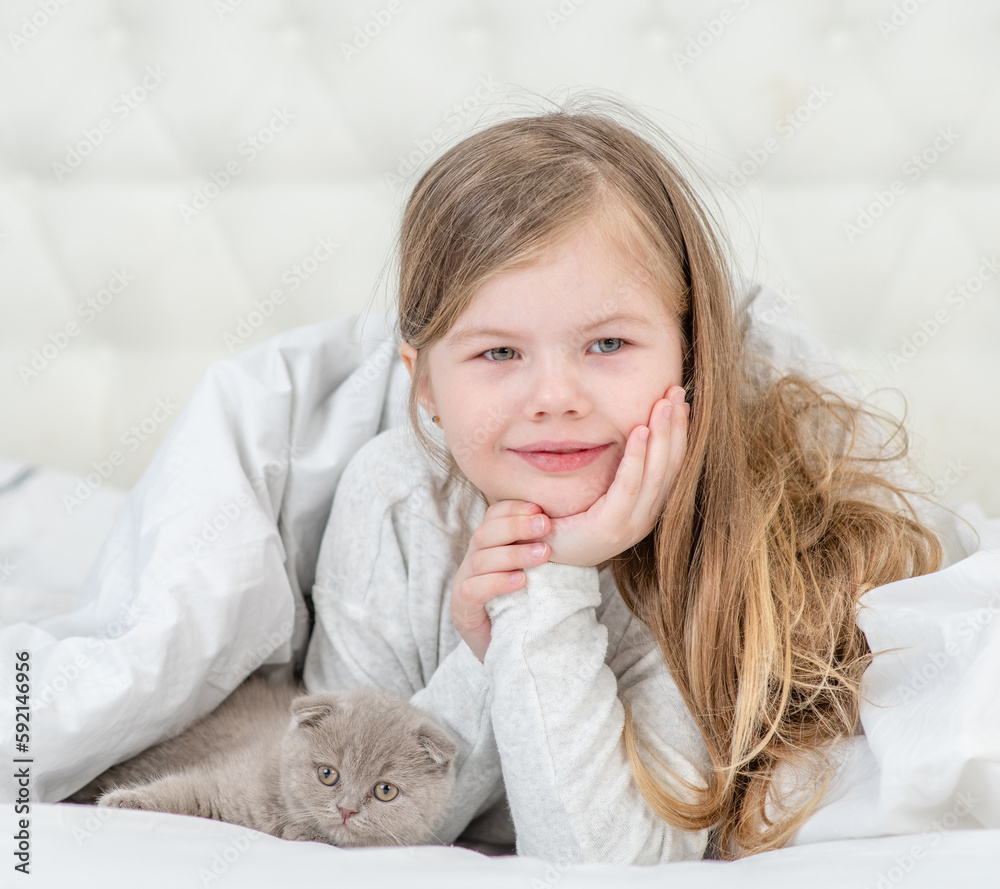 Smiling little girl and tiny kitten lying on the bed at home and looking at camera together Smiling little girl and tiny kitten lying on the bed at home and looking at camera together