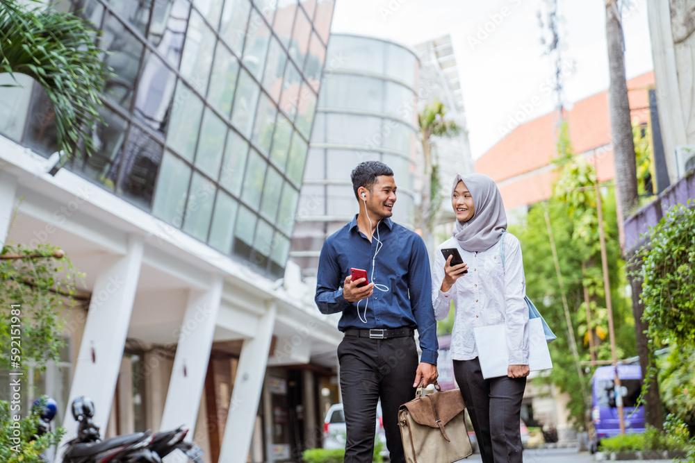 Asian male and female Muslim workers chat while walking to work together between office buildings in the city