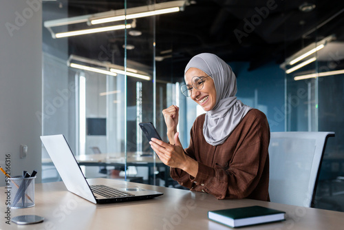 Businesswoman in hijab successful working inside office, Muslim woman at workplace holding phone, female employee received good news online, holding hand up celebrating victory and achievement
