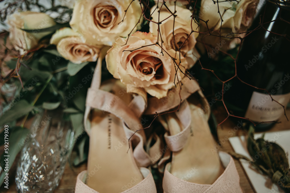 A bunch of champagne coloured roses are above a pair of strappy high heel shoes. There is a crystal wine glass and bottle in the image too.