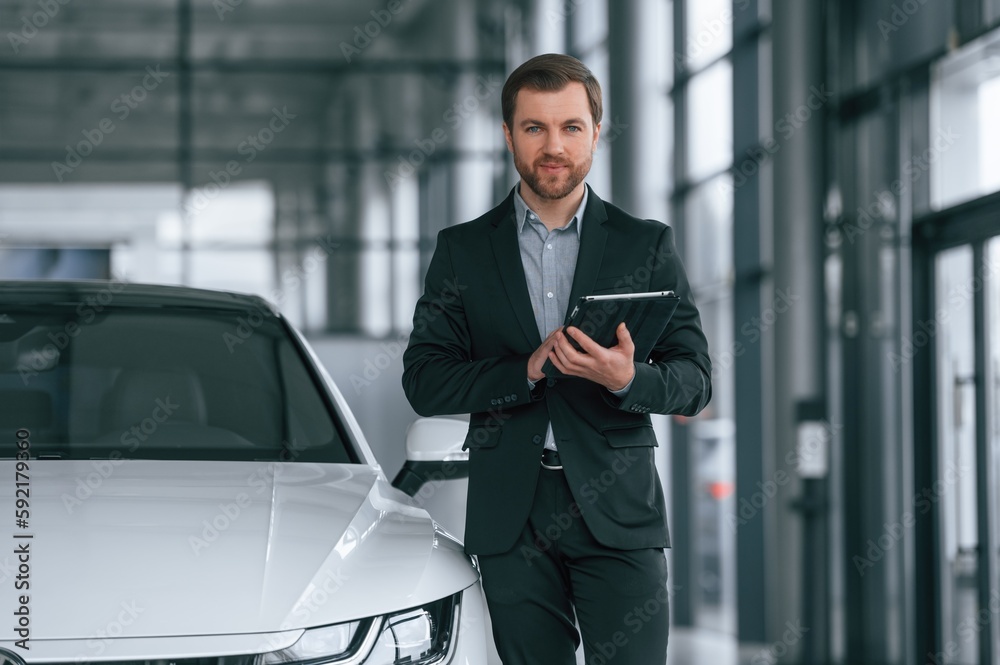With tablet in hands. Man in black suit is indoors in the car dealership near automobile With tablet in hands. Man in black suit is indoors in the car dealership near automobile