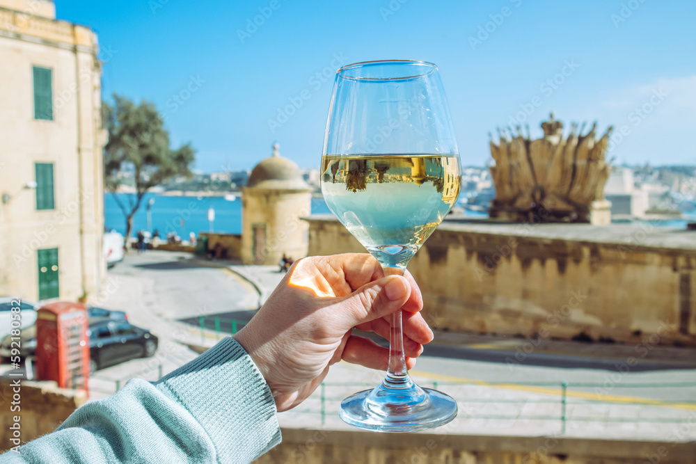Obraz na płótnie Selective focus on woman hand holding glass of white wine in beautiful city of Valletta in Malta. Background with blue sky and sea on sunny day. Wine tour concept. w salonie