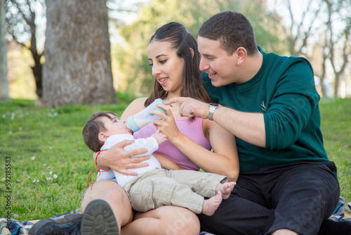 young couple happily sitting on the grass in the park while feeding their child a baby bottle © luisrojasstock