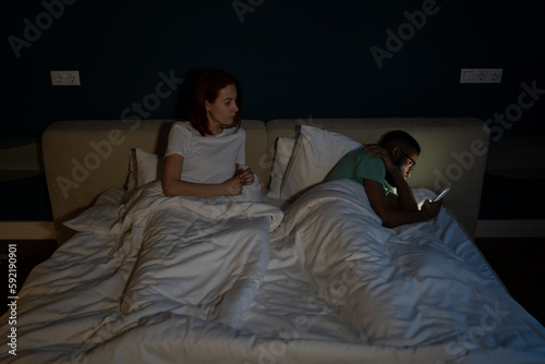 Young interracial couple lying in bed have problems in marriage life caused by smartphone addiction. Husband ignoring wife, chatting on smartphone, lost feelings for partner. Cheating in relationships