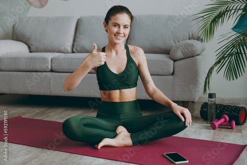 Full body young strong sporty athletic fitness trainer instructor woman wears green tracksuit sit on yoga mat show thumb up training do exercises at home gym indoor Workout sport motivation concept.