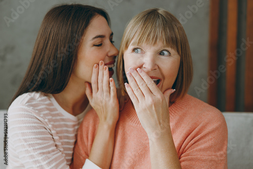 Two adult women mature mom young kid wear casual clothes whisper gossip and tells secret behind her hand hug sit on gray sofa couch stay at home flat rest relax spend free spare time. Family concept.
