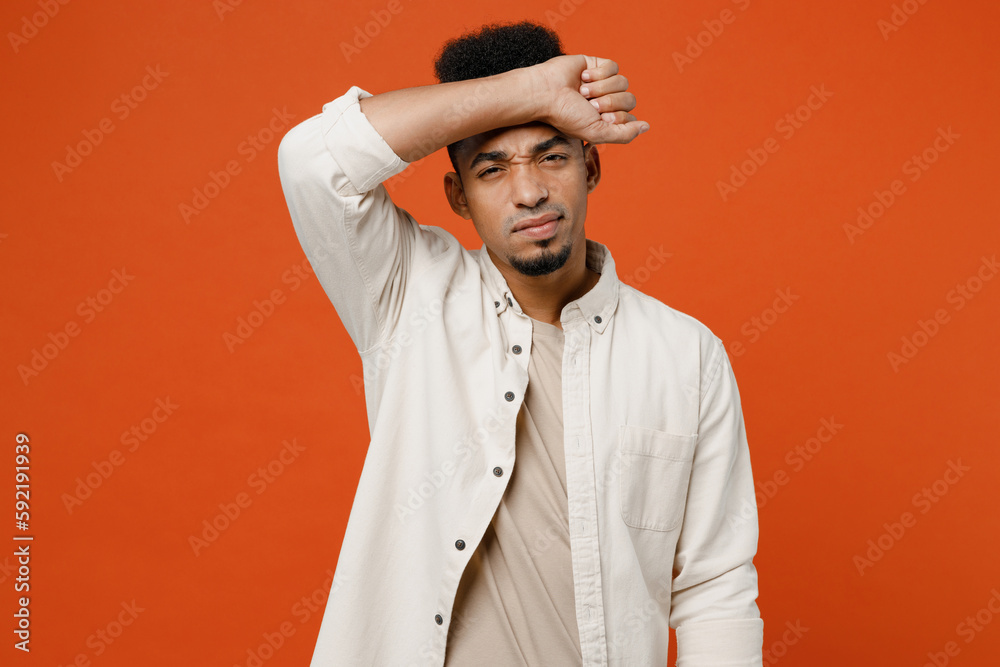 Young sad man of African American ethnicity wear light shirt casual clothes put hand on forehead look camera suffer from headache isolated on orange red background studio portrait. Lifestyle concept.