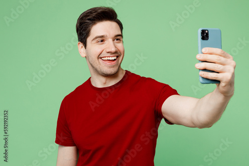 Young man he wearing red t-shirt casual clothes doing selfie shot on mobile cell phone post photo on social network isolated on plain pastel light green background studio portrait. Lifestyle concept.