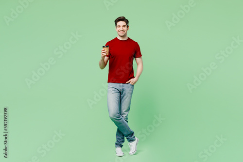 Full body young fun man he wears red t-shirt casual clothes hold takeaway delivery craft paper brown cup coffee to go isolated on plain pastel light green background studio portrait Lifestyle concept © ViDi Studio