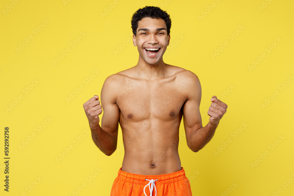 Handsome young overjoyed sexy man wear orange shorts swimsuit doing winner gesture celebrate say yes relax near hotel pool isolated on plain yellow background Summer vacation sea rest sun tan concept.