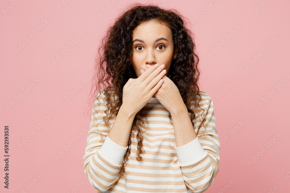 Young shocked scared fearful frightened sad woman of African American ethnicity she wear light casual clothes look camra cover mouth with hand isolated on plain pastel pink background studio portrait. Young shocked scared fearful frightened sad woman of African American ethnicity she wear light casual clothes look camra cover mouth with hand isolated on plain pastel pink background studio portrait.