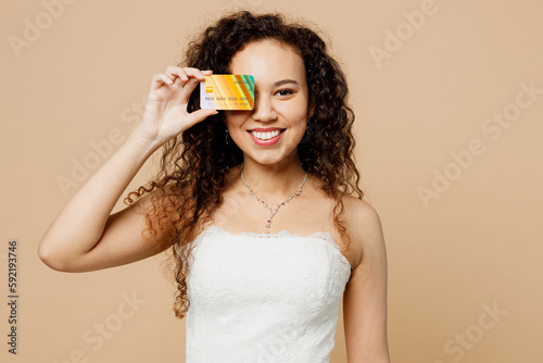 Happy young woman bride wear wedding dress posing hold cover eye with mock up of credit bank card isolated on plain pastel light beige background studio portrait. Ceremony celebration party concept.
