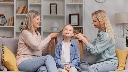 A senior woman, a grandmother, and a young mother make braids for a little blonde granddaughter on the sofa in the living room. Time spent with family, mother's day, three generations.