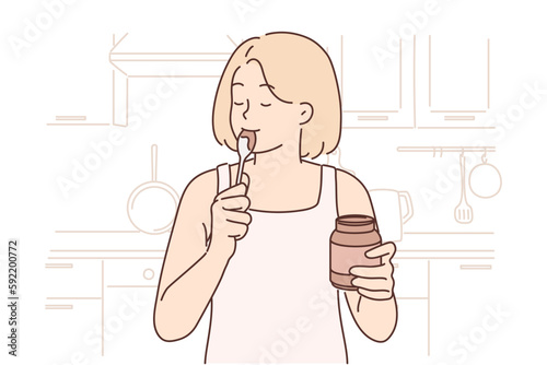Happy woman stands in kitchen with jar of chocolate paste or peanut butter and licks spoon