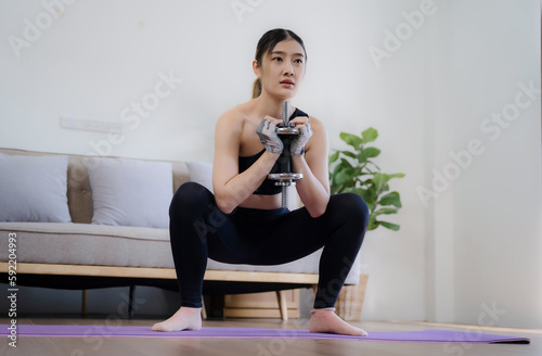Sporty woman in sportswear is sitting on the floor with dumbbells and a protein shake or a bottle of water and is using a laptop at home in the living room.