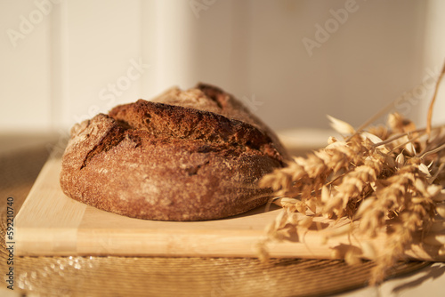 Front view of freshly baked rye bread on yeast-free starter and on mixture of whole grain, rye and wheat flour. It lies on cut board next to a dried flower with grains.Selective focus, sun.Copy space.