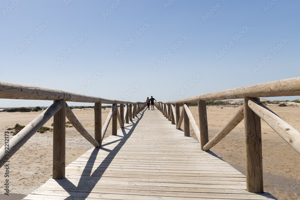 Una pareja paseando sobre una pasarela de madera en una playa del sur de Andalucía en Cádiz bajo un cielo despejado azul. Una pareja paseando sobre una pasarela de madera en una playa del sur de Andalucía en Cádiz bajo un cielo despejado azul.