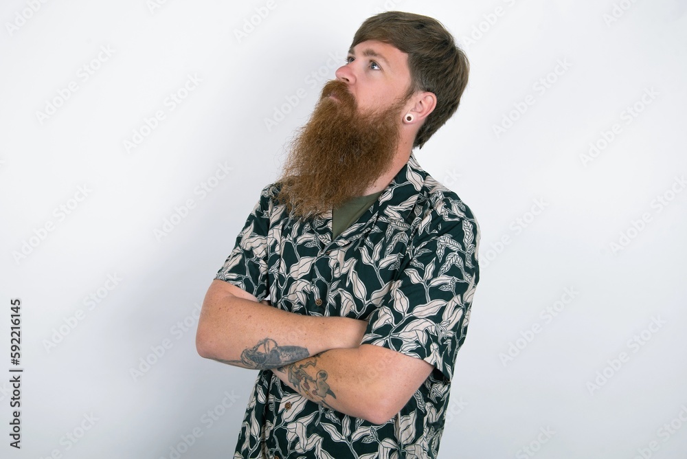 Charming thoughtful Red haired man wearing printed shirt over white studio background stands with arms folded concentrated somewhere with pensive expression thinks what to do Charming thoughtful Red haired man wearing printed shirt over white studio background stands with arms folded concentrated somewhere with pensive expression thinks what to do