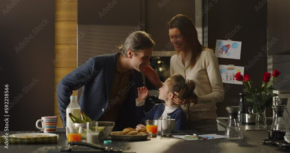 Happy Family Portrait: Father Kissing Mother and Daughter Before Leaving for Work in the Morning. Little Girl Having Breakfast While her Mother Gets her Ready for School Happy Family Portrait: Father Kissing Mother and Daughter Before Leaving for Work in the Morning. Little Girl Having Breakfast While her Mother Gets her Ready for School