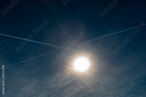 Airplane with Vapor Trails in Cross Symbol Against Sun with Blue Sky in Ticino, Switzerland.