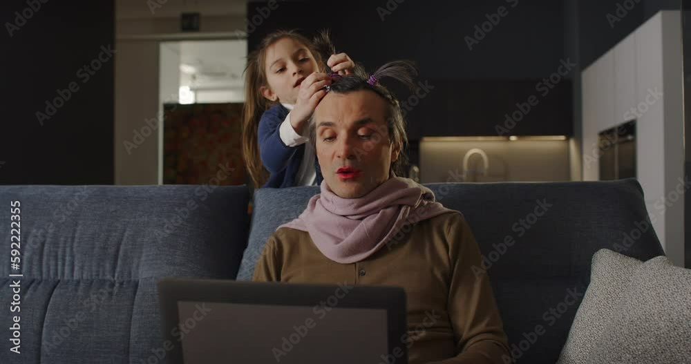 Funny Family Situation: Portrait of a Man Having a Video Call Conference with his Colleagues Using Laptop Computer at Home While his Daughter is Playing Makeup on him. Work from Home Concept
