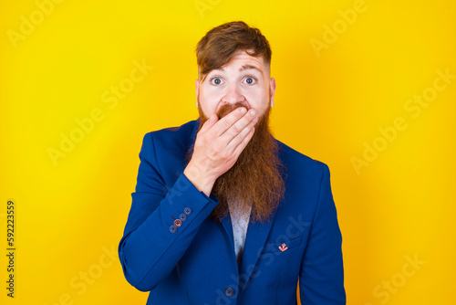 Emotional red haired man wearing blue suit over yellow studio background gasps from astonishment, covers opened mouth with palm, looks shocked at camera. Reaction concept