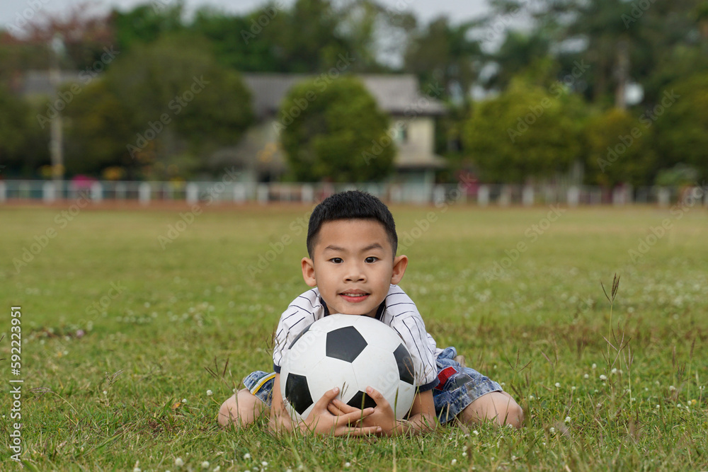 Asian boy sits with his chin on a soccer ball in the grass, in the concept of outdoor activities, sports, playgrounds, leisure activities. Soft and selective focus. Asian boy sits with his chin on a soccer ball in the grass, in the concept of outdoor activities, sports, playgrounds, leisure activities. Soft and selective focus.