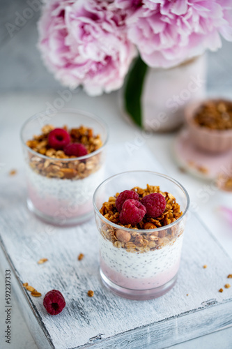  Raspberry chia seed parfait made with granola and berries in a cups with peony flowers on the background.