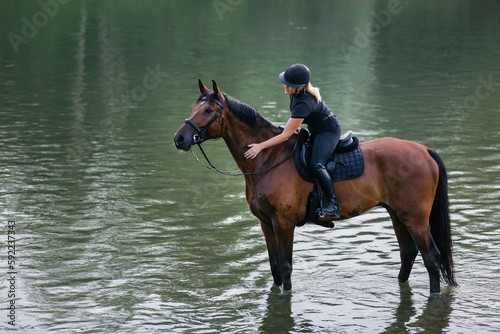 Female horseback rider in a black jockey outfit riding a chestnut horse along the river at sunset. Recreation, equitation, and nature concept.