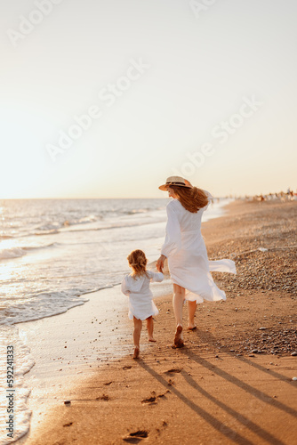 A little girl and a young mother in white dresses and a hat walk barefoot on the sand on the ocean at sunset. Wind blows hair