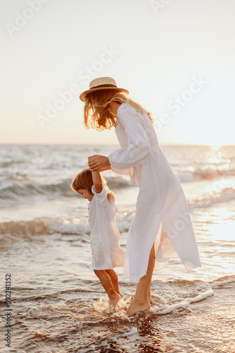 A young girl, mother, with a little daughter in white dresses, walk on the water on the seashore at sunset. The sun breaks through your hair