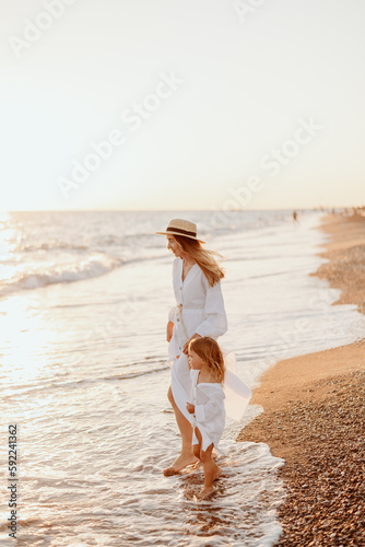 A young girl, mother, with a little daughter in white dresses, walk on the water on the seashore at sunset.