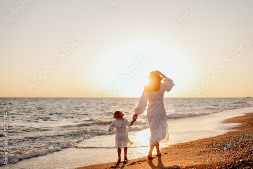 A young mother holds her little daughter by the hand and together they walk along the ocean towards the sunset. Girls in white dresses and long hair