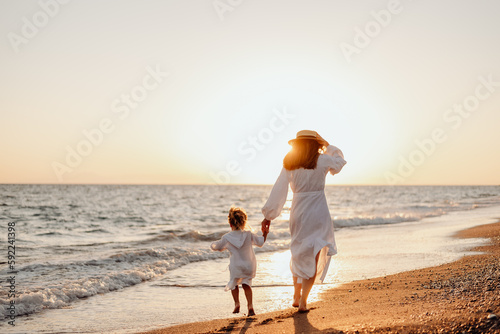 A young mother holds her little daughter by the hand and together they run along the ocean towards the sunset. Girls in white dresses and long hair