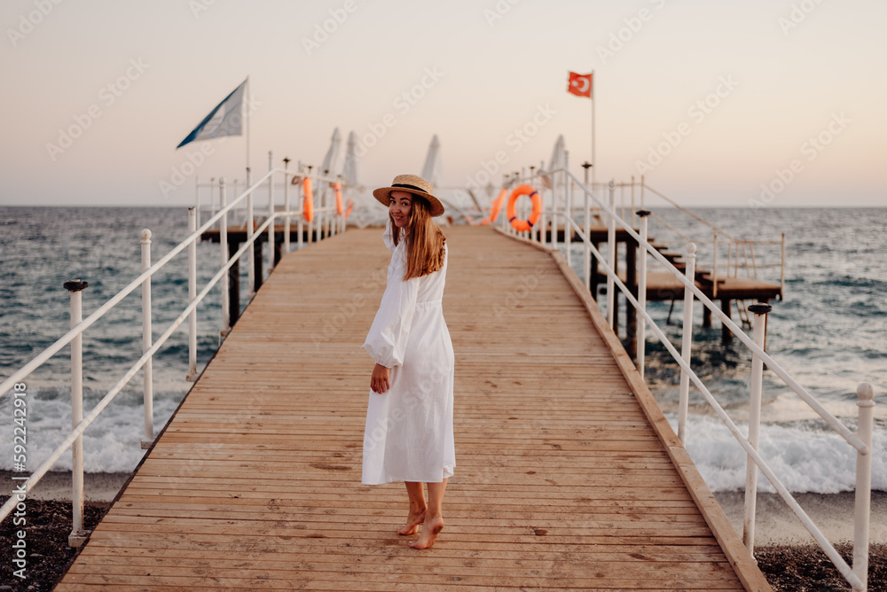 A young girl with long hair in a white dress and a straw hat walks towards the ocean along a wooden pier at sunset