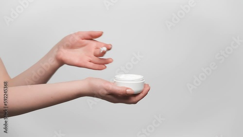 Woman is holding jar of face cream in her hand, taking a sample with a finger and applying it on a hand, gray background, copy space on the right. 4k, slow motion.