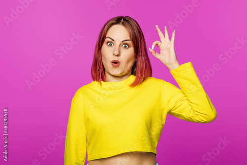 Portrait of happy and positive woman with pink hair looking amazed and shows ok sign, standing in yellow t-shirt on pink background