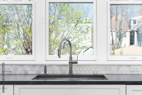A kitchen sink detail with white cabinets  grey countertop  a marble hexagon backsplash  and large  bright windows.