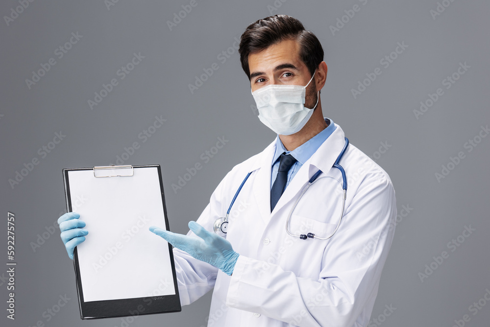 A male doctor in a white coat and a medical mask with a notepad in his hand looks at the camera on a gray isolated background, copy space, space for text A male doctor in a white coat and a medical mask with a notepad in his hand looks at the camera on a gray isolated background, copy space, space for text