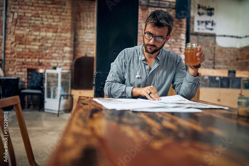 Handsome young man with glasses holding drink and reading newspaper in modern cafe.