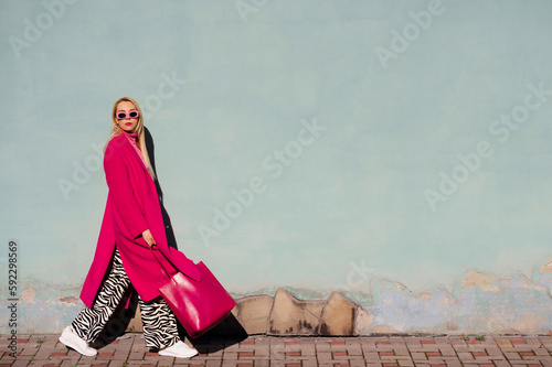 Foto Fashionable confident woman wearing trendy pink sunglasses, fuchsia color midi coat, zebra print trousers, white sneakers, holding tote, shopper bag, walking outdoors