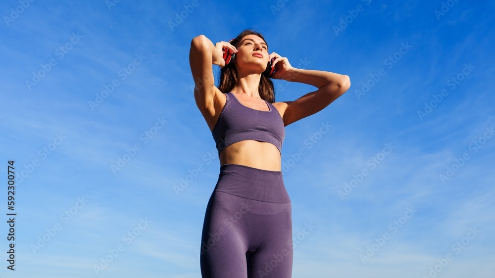 A young woman puts on headphones before training outside against a blue sky background A young woman puts on headphones before training outside against a blue sky background