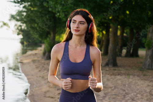 A brunette in a tracksuit runs around on the beach with headphones. Morning workout in summer