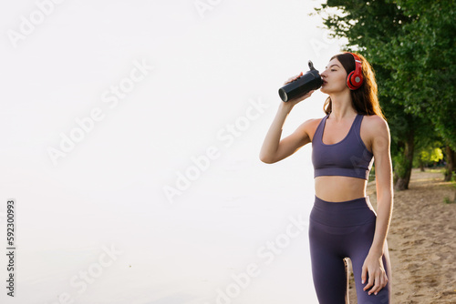 A sporty woman drinks water after jogging along the sea. A model in a tracksuit poses outside