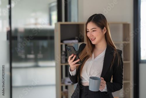 Portrait of businesswoman holding coffee cup and using a mobile phone  standing and smiling  at an office.