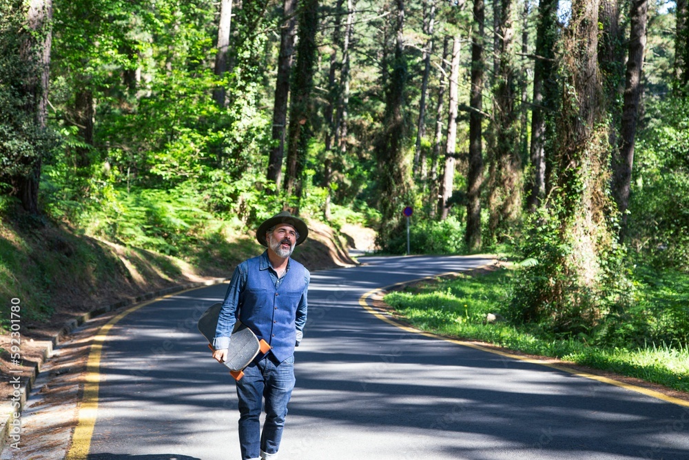 View of a man carrying a skateboard in his hand while walking on the road through a forest View of a man carrying a skateboard in his hand while walking on the road through a forest