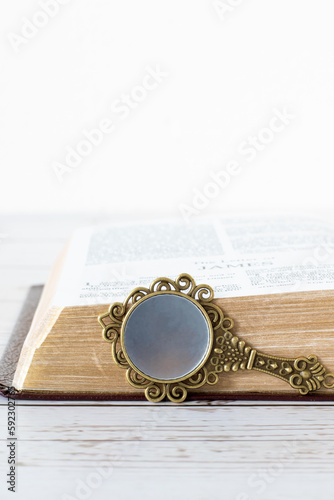 Antique mirror in front of an open holy bible book with golden pages isolated on wooden table with white background. Vertical shot, a closeup. Spiritual reflection and obedience, Christian concept.